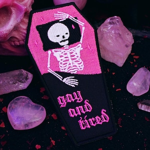 Könnte beinhalten: Rosa-schwarzer gestickter Bügel-Patch mit einem Skelett in einem Sarg mit dem Text "gay and tired".