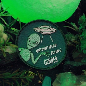May include: A green embroidered patch with a cartoon alien holding a flying saucer. The text on the patch reads "Unidentified Flying Gender".