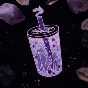 Puede incluir: Un parche bordado que representa una taza de boba tea en morado y blanco, con un pentagrama en la tapa y cristales en su interior. El fondo es negro con cristales morados dispersos.