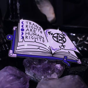 May include: A white and purple enamel pin in the shape of an open book. The book has the text "TRANS RIGHTS ARE HUMAN RIGHTS" and a pentagram symbol. The pin features a purple border and moon phases.