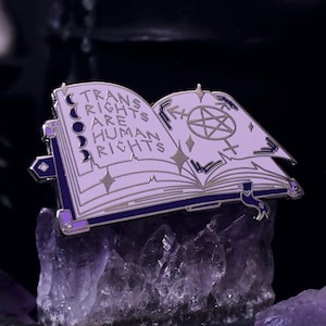 Op de afbeelding: Een lavendelkleurige emaille pin in de vorm van een open boek. De pin heeft de tekst "TRANS RIGHTS ARE HUMAN RIGHTS" en is versierd met manen, sterren en een pentagram. De pin heeft een zilveren rand en paarse accenten.