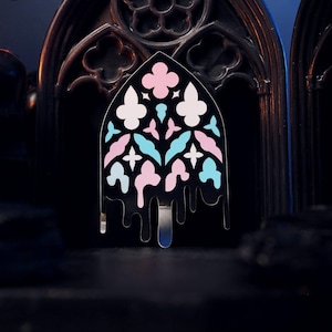 May include: A black enamel pin shaped like a pointed gothic arch window. The design features a symmetrical pattern of light pink, white, and light blue shapes. The bottom edge of the pin has a dripping effect, with black metal extending downward.