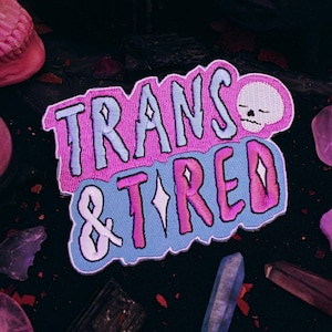 May include: A blue and pink embroidered patch with the text "Trans & Tired" and a small white skull with black eyes.