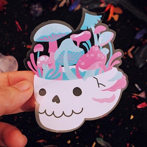 May include: A whimsical sticker featuring a white skull with a cheerful expression, its head filled with colorful mushrooms in shades of pink and blue. The sticker is held against a dark, textured background.