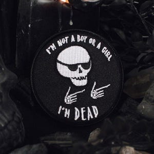 May include: Black and white embroidered patch with a skull wearing sunglasses and the text "I'm not a boy or a girl I'm dead".