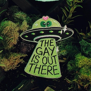 May include: Green and black embroidered patch of a UFO with the text "The Gay is Out There" in a green font.