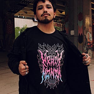 Puede incluir: Una persona lleva una chaqueta negra sobre una camiseta negra con un gráfico rosa y azul de una cabeza de lobo con llamas. El gráfico tiene el texto "Wrath" y "Flames" en una fuente estilizada.