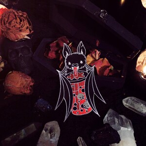 May include: A black and white enamel pin of a bat drinking a red and white boba tea. The bat has a red tongue sticking out and the boba tea has white boba pearls inside. The pin is surrounded by dried roses, crystals, and candles.