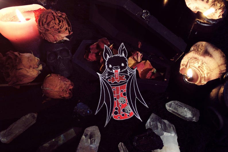 May include: A black and white enamel pin of a bat drinking a red and white boba tea. The bat has a red tongue sticking out and the boba tea has white boba pearls inside. The pin is surrounded by dried roses, crystals, and candles.