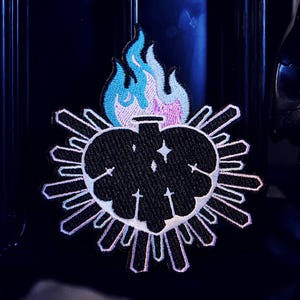 May include: Embroidered patch featuring a black heart with the word "SICK" in white, surrounded by a white outline. Above the heart is a blue and pink flame design. The patch is set against a dark background.