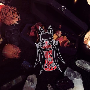 May include: A black bat with white wings is drinking from a red and white cup with black polka dots. The bat is surrounded by dried roses, crystals, and candles.