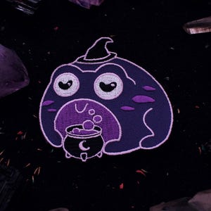 May include: Purple embroidered patch of a frog wearing a witch's hat and holding a cauldron with bubbling potion.