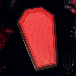 Könnte beinhalten: Ein roter Sarg-Notizblock mit horizontalen Linien und einer Fledermaus-Illustration. Der Notizblock ist von roten Rosen und dunklen, gotischen Elementen auf schwarzem Hintergrund umgeben.