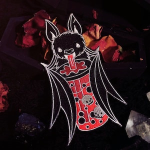 May include: A black and white embroidered patch of a cartoon bat drinking a red and white boba tea. The bat has large, white eyes and a wide, toothy grin. The boba tea has black boba pearls and a red straw.