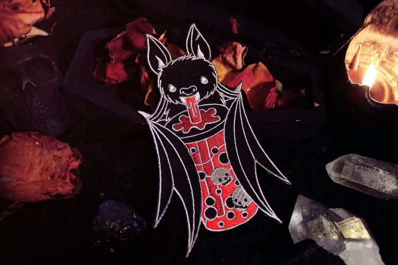 May include: A black and white embroidered patch of a cartoon bat drinking a red and white boba tea. The bat has large, white eyes and a wide, toothy grin. The boba tea has black boba pearls and a red straw.