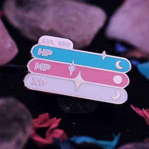 May include: A white, pink, and blue enamel pin with a game-like interface. The pin reads "LVL 100", "HP", "MP", and "XP".