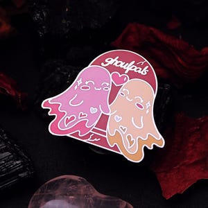 May include: A pink and orange enamel pin featuring two cartoon ghosts with hearts and stars. The text "ghoul pals" is written in a white arch above the ghosts.