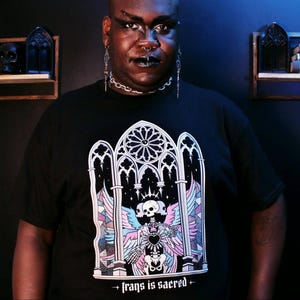 Peut inclure: T-shirt noir avec un motif gothique représentant un crâne, des ailes et le texte "trans is sacred". Le motif intègre des couleurs pastel. La personne sur l'image porte des bijoux argentés.