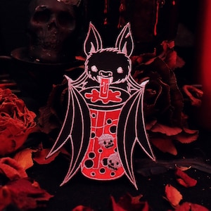 May include: A black bat with white accents is drinking from a red cup with white skulls and bubbles. The bat is holding the cup with its wings. The background is a dark red with roses.