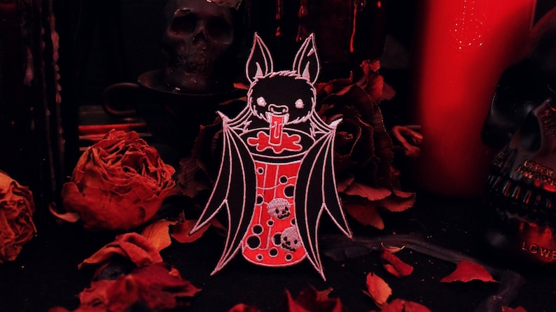 May include: A black bat with white accents is drinking from a red cup with white skulls and bubbles. The bat is holding the cup with its wings. The background is a dark red with roses.