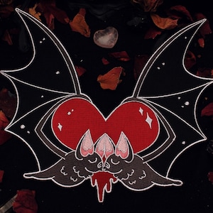 May include: A black and red bat-themed design featuring a heart shape with bat wings and two kissing bats. The design has white outlines and small star details. The image has a dark background with scattered dried leaves.