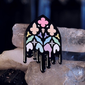 May include: A black enamel pin, shaped like a stained-glass window, with a colourful floral design in pink, yellow, blue, and white. The pin has a dripping effect at the bottom and is displayed on crystals.