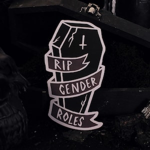 Puede incluir: Una pegatina en blanco y negro de un ataúd con las palabras "RIP Gender Roles" en ella. La pegatina está sobre un fondo oscuro.