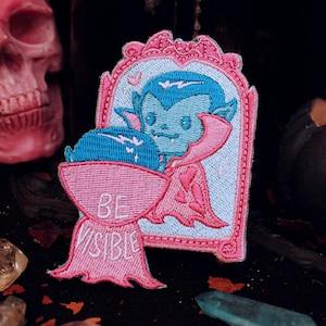 Può includere: Toppa ricamata con un vampiro dei cartoni animati in una cornice rosa, con la scritta "BE VISIBLE". Il vampiro ha i capelli blu e un mantello rosa. La toppa è ambientata su uno sfondo scuro con un teschio e cristalli.