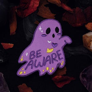Może przedstawiać: Fioletowy duch z przyjazną twarzą i napisem "BE AWARE" w żółtych literach. Duch jest otoczony jesiennymi liśćmi i kryształami.