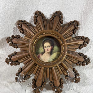 May include: A gold sunburst frame with a miniature portrait of a woman in a white dress and blue headscarf. The frame has a decorative design with a raised edge.