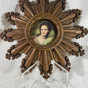 May include: A miniature portrait of a woman in a gold frame with a sunburst design. The frame is made of a gold-coloured metal and has a raised design. The portrait is of a woman with dark hair and a white dress. The background of the portrait is a light blue.