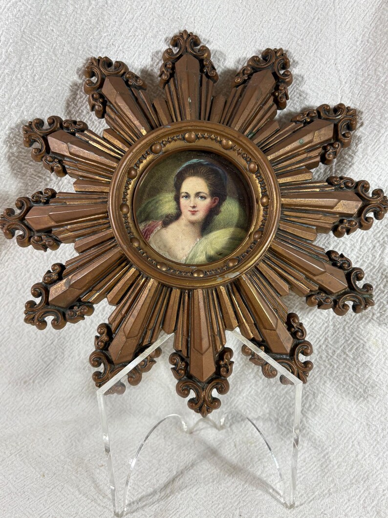 May include: A miniature portrait of a woman in a gold frame with a sunburst design. The frame is made of a gold-coloured metal and has a raised design. The portrait is of a woman with dark hair and a white dress. The background of the portrait is a light blue.