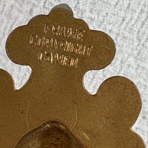 May include: A gold-coloured metal ornament with a decorative design. The ornament has the text "FRAME" and "COPYRIGHT" on it.