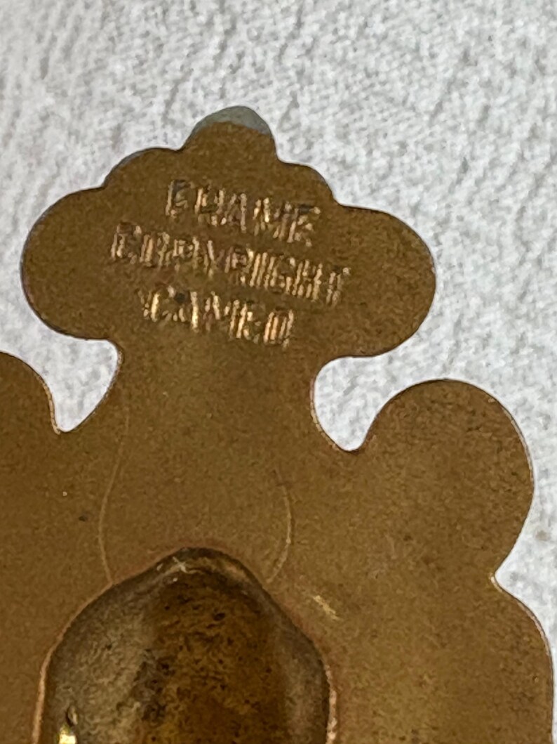 May include: A gold-coloured metal ornament with a decorative design. The ornament has the text "FRAME" and "COPYRIGHT" on it.
