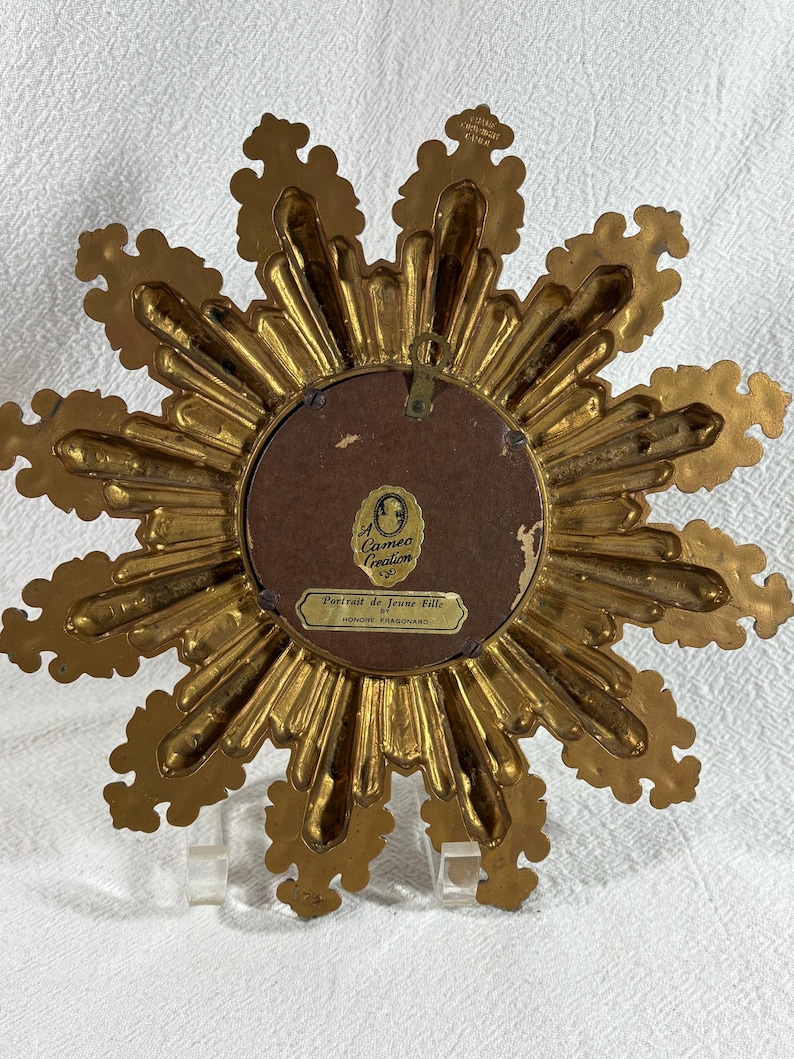 May include: Gold-toned starburst-shaped picture frame with a cameo creation label on the back. The label reads "Cameo Creation" and "Portrait de Jeune Fille par Honore Fragonard".