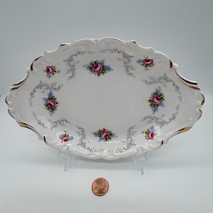 Puede incluir: Plato ovalado blanco para servir con borde dorado y un patrón floral de rosas rosas y azules. El plato tiene un borde festoneado y un delicado diseño plateado.