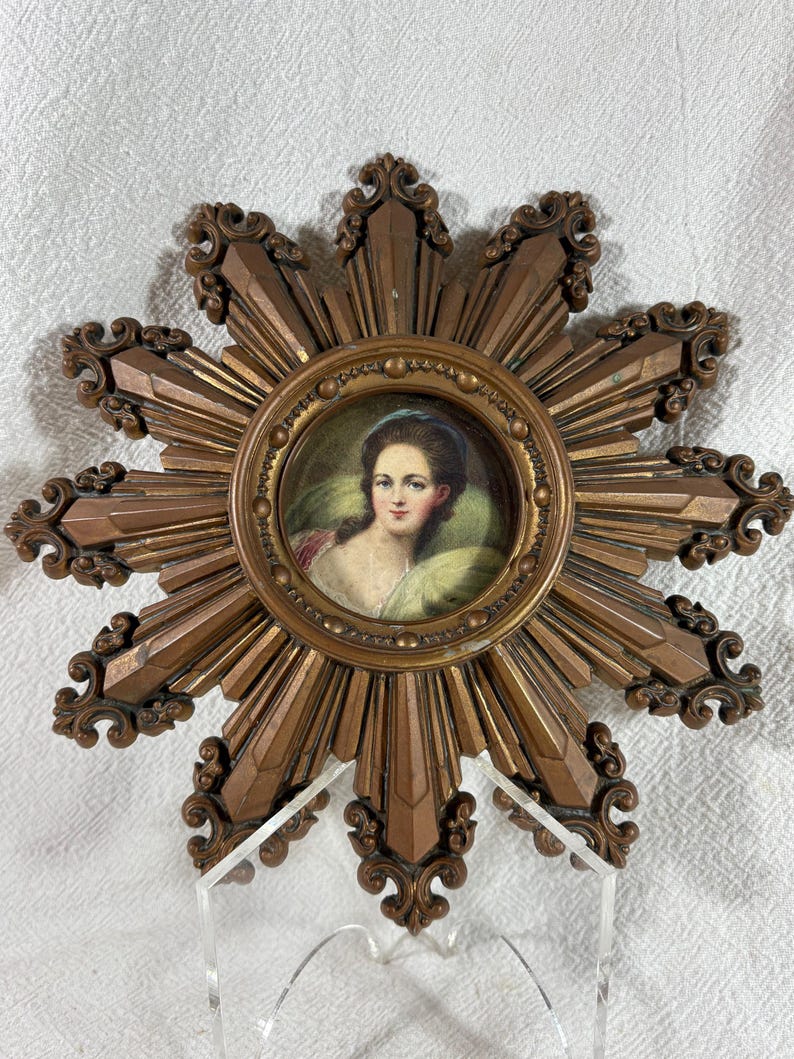 May include: A gold sunburst frame with a miniature portrait of a woman in a green dress. The frame has a decorative design with a raised edge and a beaded border.