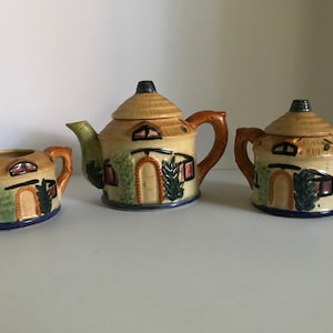 Puede incluir: Un juego de té de cerámica vintage con un diseño de cabaña. La tetera, la jarra de crema y el azucarero están decorados con una escena de cabaña marrón, verde y azul.