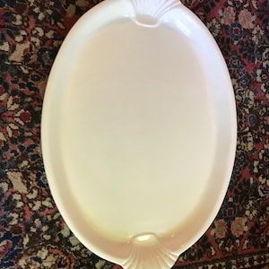 May include: An oval, white serving platter with shell-shaped handles. The platter has a smooth, glossy surface and is designed for serving food. The handles add a decorative touch to the serving piece. This platter is suitable for various occasions.