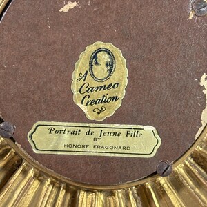 May include: An ornate gold-coloured frame with a brown background. The frame has a label that reads "Portrait de Jeune Fille by Honore Fragonard". There is a smaller label that reads "A Cameo Creation".
