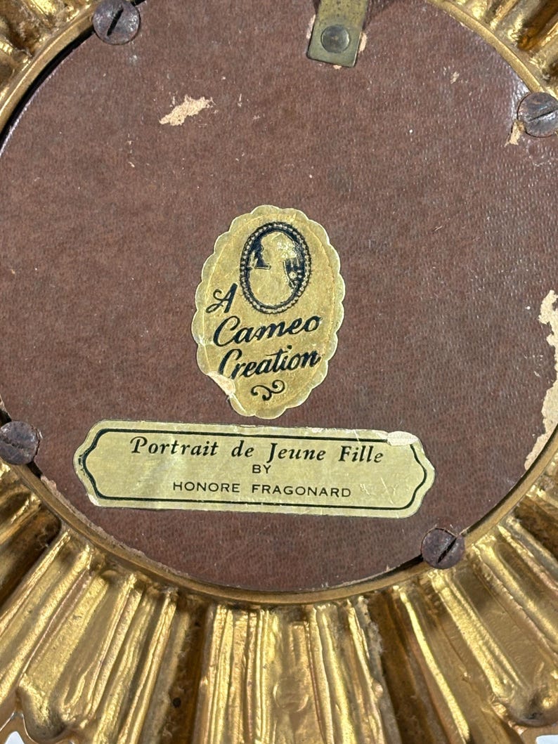 May include: An ornate gold-coloured frame with a brown background. The frame has a label that reads "Portrait de Jeune Fille by Honore Fragonard". There is a smaller label that reads "A Cameo Creation".