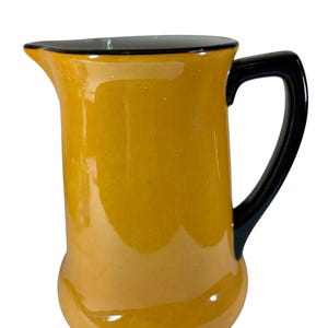 May include: A ceramic pitcher with a rounded base and a black handle. The pitcher is a warm yellow color with a black rim. The spout is designed for easy pouring. This pitcher is suitable for serving beverages.