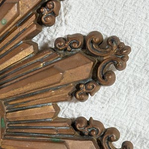 May include: Close-up of a gold-toned metal sunburst design with intricate details. The sunburst has a raised, textured surface and a slightly aged appearance.