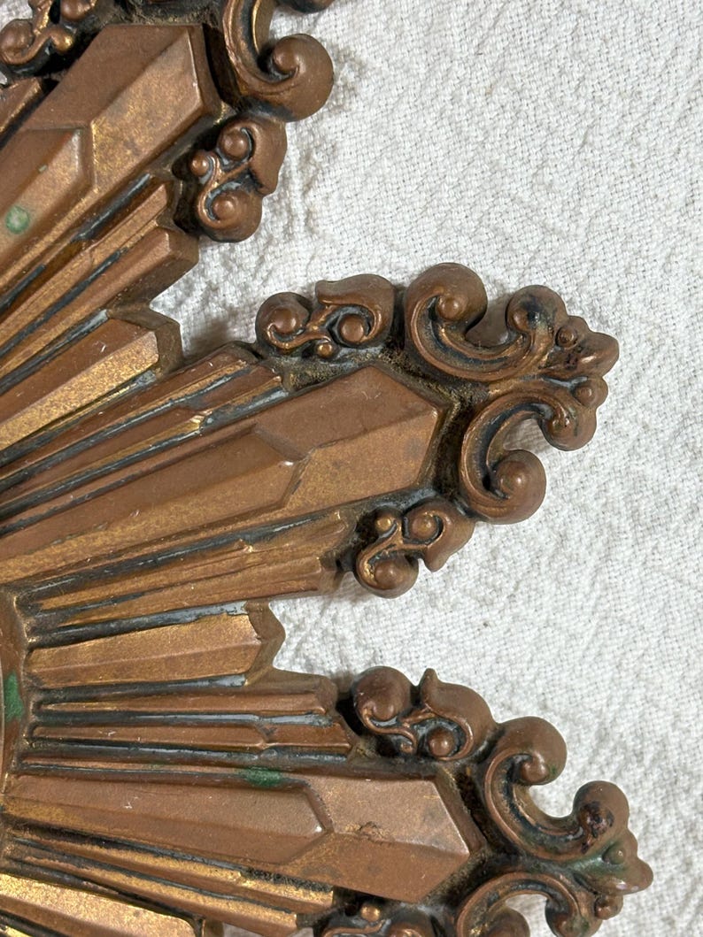 May include: Close-up of a gold-toned metal sunburst design with intricate details. The sunburst has a raised, textured surface and a slightly aged appearance.