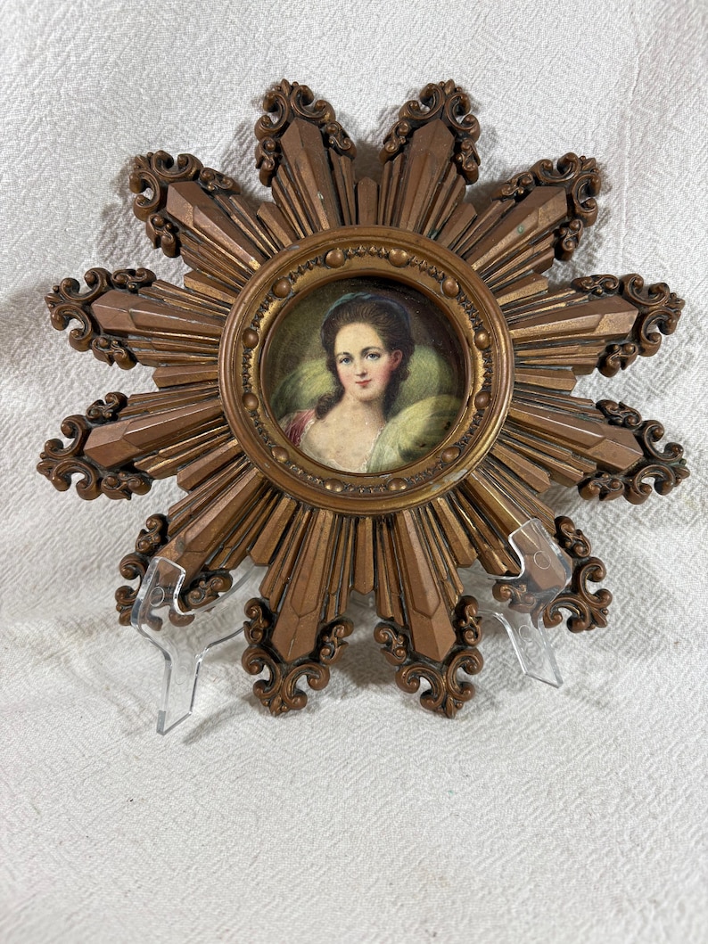 May include: A gold sunburst frame with a miniature portrait of a woman in a white dress and blue headscarf. The frame has a decorative design with a raised edge.
