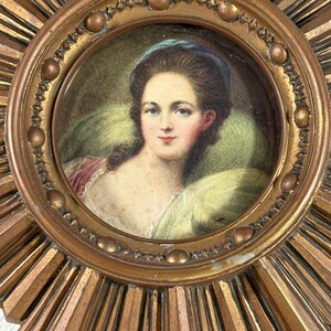 May include: A gold sunburst mirror with a round, framed portrait of a woman in a blue headband. The portrait is painted in watercolour and features a woman with brown hair and a white dress.