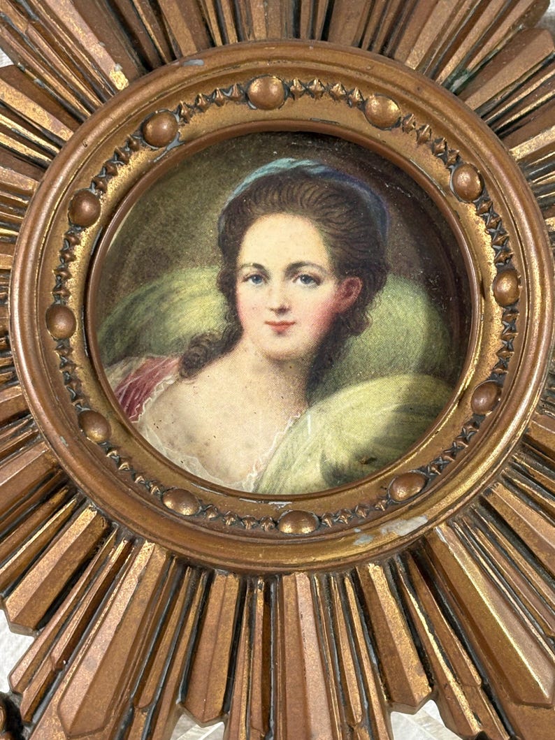 May include: A gold sunburst mirror with a round, framed portrait of a woman in a blue headband. The portrait is painted in watercolour and features a woman with brown hair and a white dress.