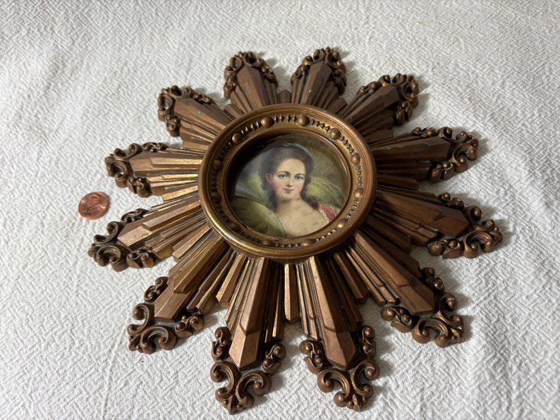May include: A gold sunburst wall decor with a round portrait of a woman in a white dress and a gold frame.
