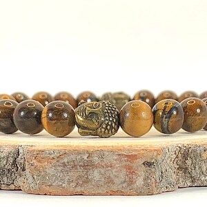 May include: A brown tiger's eye beaded bracelet with a gold Buddha head charm. The bracelet is on a wooden surface.