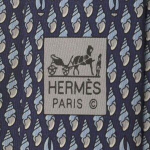 May include: A dark blue necktie featuring a repeating pattern of light blue and white spiral seashells. A rectangular gray label is centered on the fabric, displaying a black graphic of a horse-drawn carriage and the text HERMÈS PARIS ©.